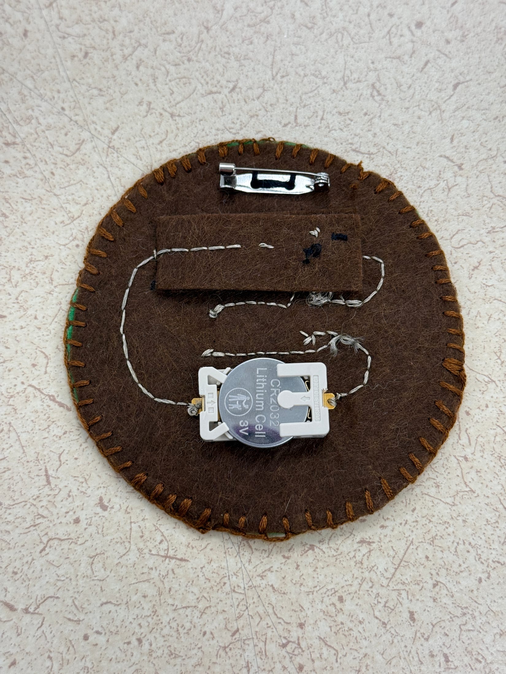 Back of the cat badge showing conductive thread, a battery, and a pin